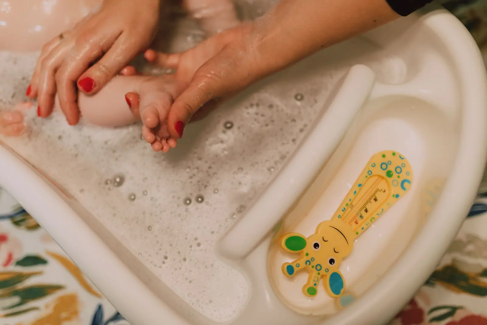 Adult hands supporting a baby's legs in a small white tub beside a giraffe-shaped bath thermometer