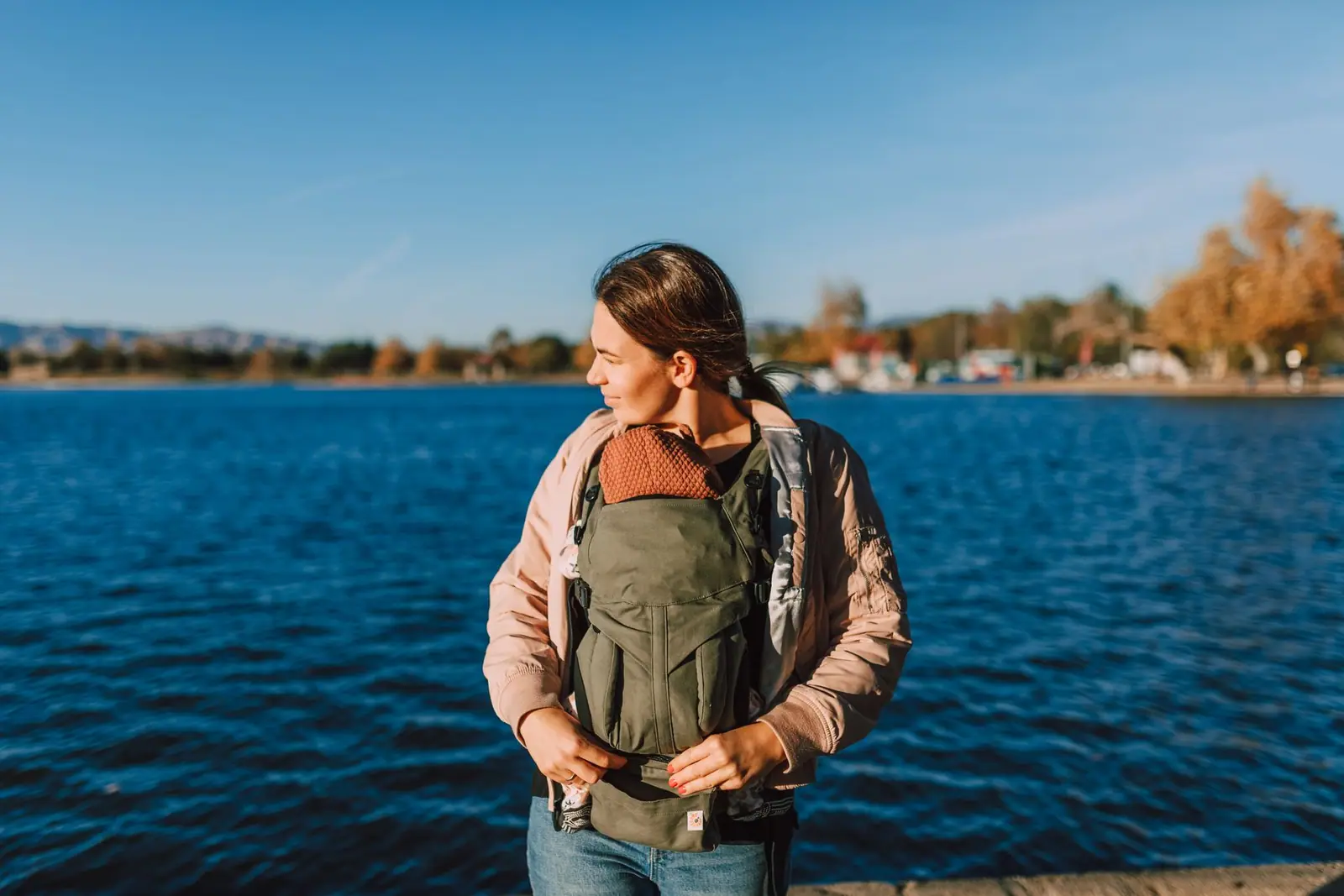 A woman wearing an olive-green baby carrier beside a blue lake