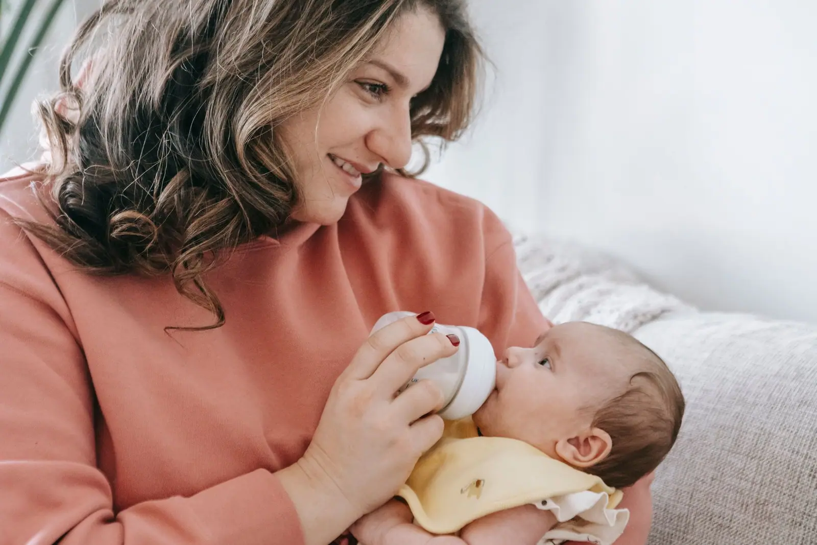 An adult in a pink shirt seated on a sofa while feeding a baby