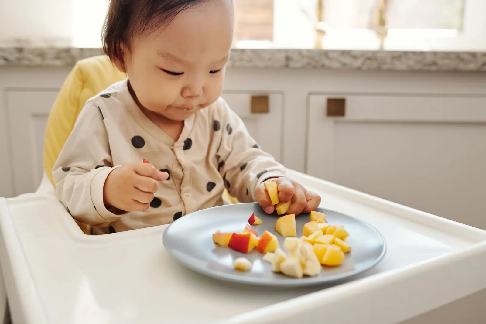 A baby in a high chair reaching for pieces of fruit on a plate